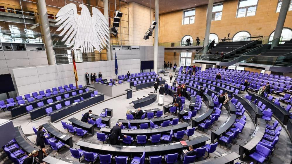 Inside the Bundestag, where there are many purple chairs in a semi-circle, and a huge eagle - the logo of the Bundestag  - hanging above the speaker. There are a few people milling around but it's mainly empty.