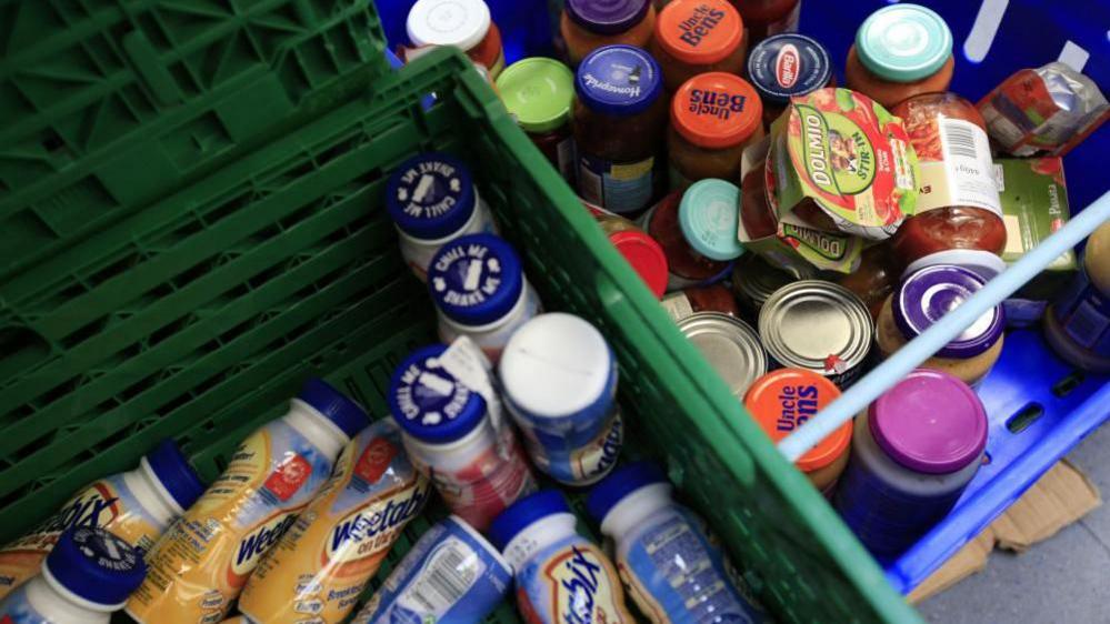 A variety of containers of breakfast drinks, jars of sauce and tinned goods in two green plastic baskets at a food bank