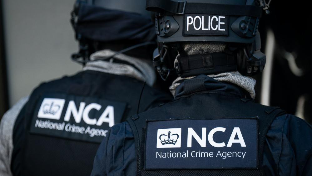 Two National Crime Agency (NCA) officers pictured from behind. They are wearing black jackets and NCA bulletproof vests, as well as black helmets.