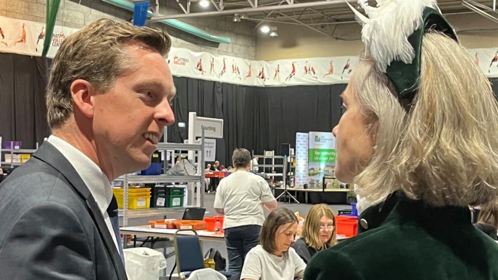 Tom Pursglove with short fair hair wearing a grey jacket and talking to a woman in black and white High Sherriff uniform.  An election count is underway in the background.