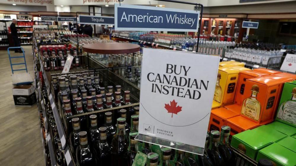 A sign that reads ''Buy Canadian Instead'' is displayed on top of bottles, hanging above another sign that reads "American Whiskey"