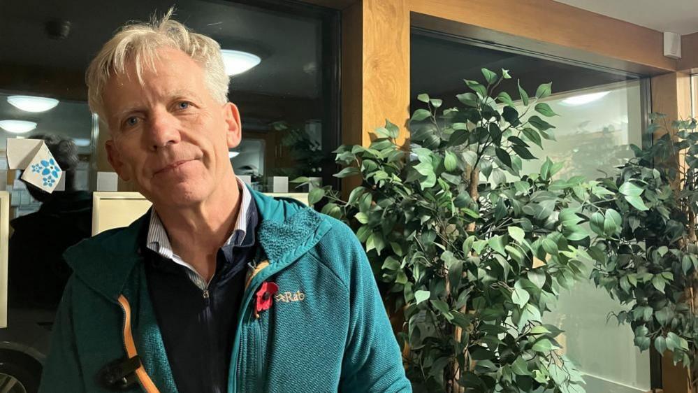 Richard Darn stands in the lobby of Husthwaite Village Hall. He has short white hair and wears a blue jacket with a Remembrance poppy on the chest , a navy sweater and a striped shirt. In the background, a large potted plant can be seen.