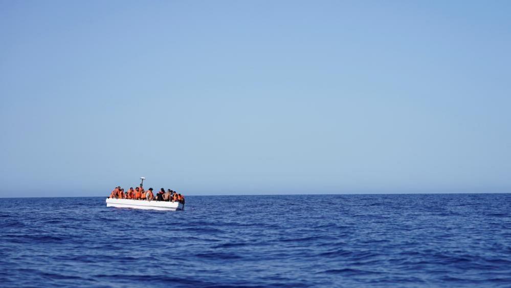 A vast open blue ocean and sky with a small white boat packed with people in orange lifevests