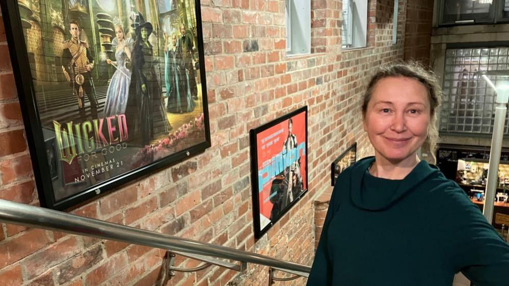 Cath Sharp stands on the stairs at the City Screen Picturehouse in York, next to a poster for Wicked: For Good.