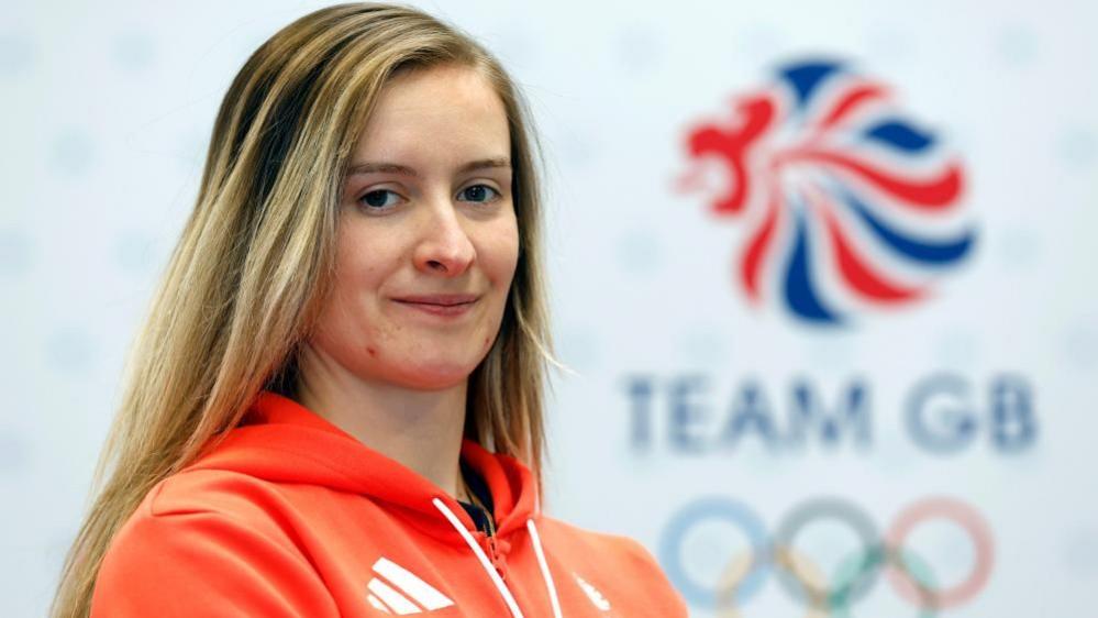 Charlotte Worthington with long brown hair wearing an orange Team GB fleece and standing in front of a Team GB sign with the five Olympic rings underneath.