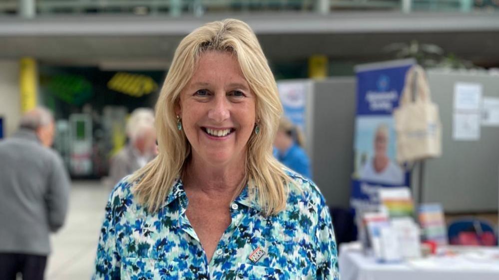 Zoe Bellingham is standing inside The Forum in Norwich at an event where display hoardings are visible in soft focus. She is wearing a highly patterned blue, black and white blouse, and she has below the shoulder length blonde hair. She is smiling.