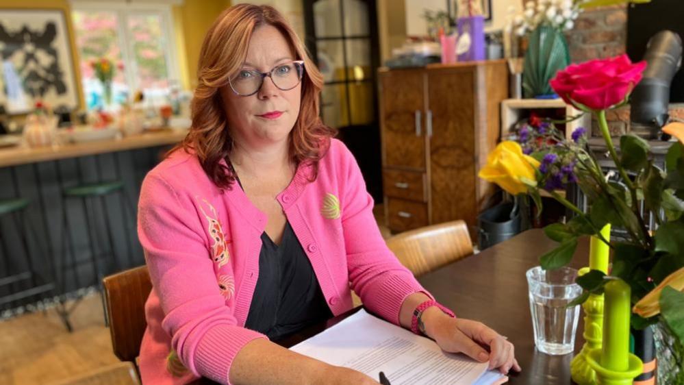 A woman with long wavy hair wears a pink cardigan and glasses, and looks into the camera. She is sitting at a dining table with papers and a glass of water in front of her, and a kitchen behind her.