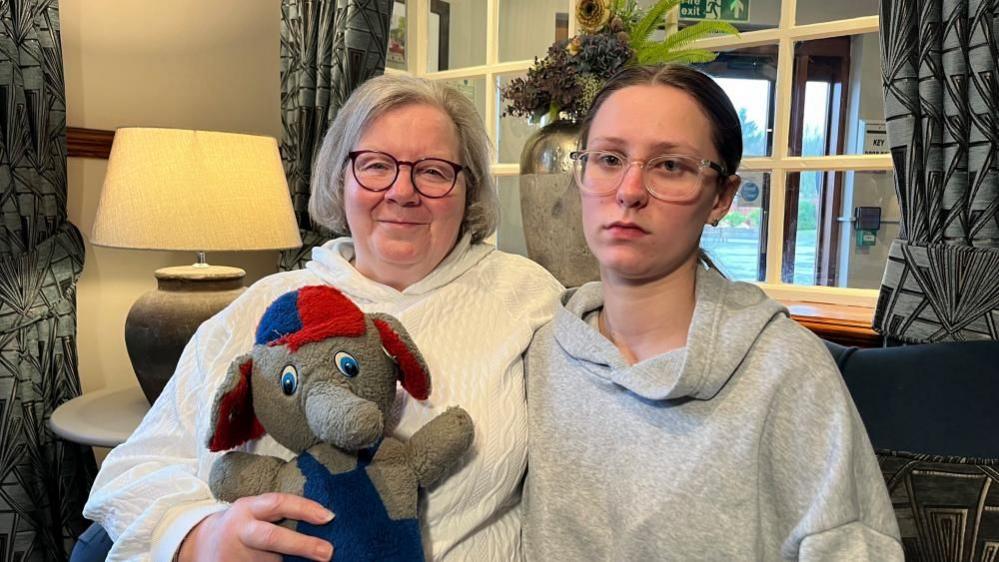 Helen Thrower, Terry McSpadden's mum, has silvery hair at shoulder length. She's wearing glasses and a white rope-knitted jersey. She's holding Terry's toy elephant from when he was a child; it is grey, has blue dungarees and a red and blue cap on its head. Terry's daughter Kayce (pronounced Casey) has long brown hair; tied back in a ponytail. She is wearing a grey hoodie, and glasses. They are sitting in a hotel reception.