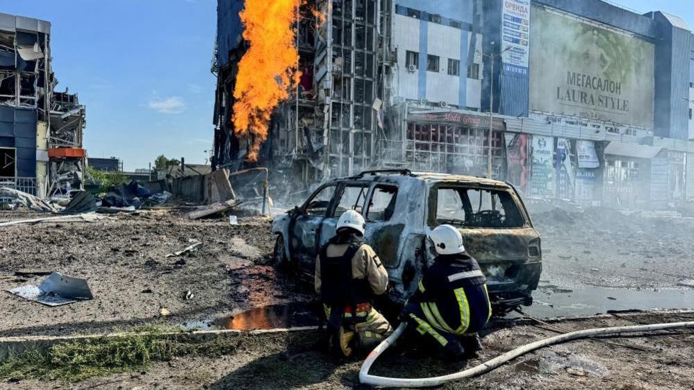 A badly damaged supermarket building, a flame is visible from what appears to be a burning gas line. Two firefighters are crouching next to a destroyed car and appear to be trying to extinguish it.