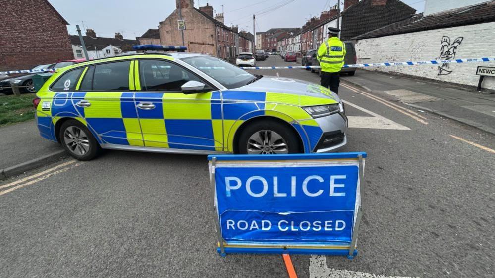 A yellow and blue police car parked in a road with a "police road closed" sign in the foreground and a police officer behind the car. There is police tape across the road and terraced houses are visible beyond.