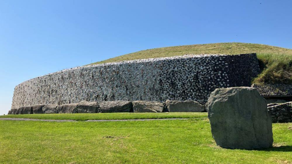 Newgrange tomb, a mound built with grey stones on top of which is grass. Large boulders sit in front of this