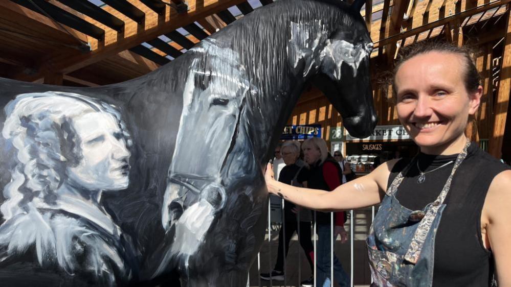 Lois Cordelia smiling, next to her artwork; a black horse painted with images of Black Beauty and author Anna Sewell.