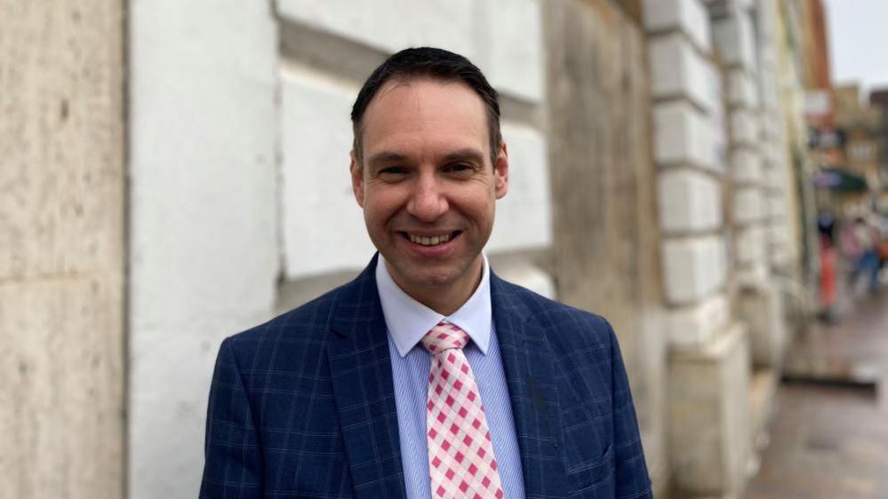 Dan Lister with short dark hair wearing a blue jacket over a blue shirt, and a red and white tie. He is standing in front of a stone wall.