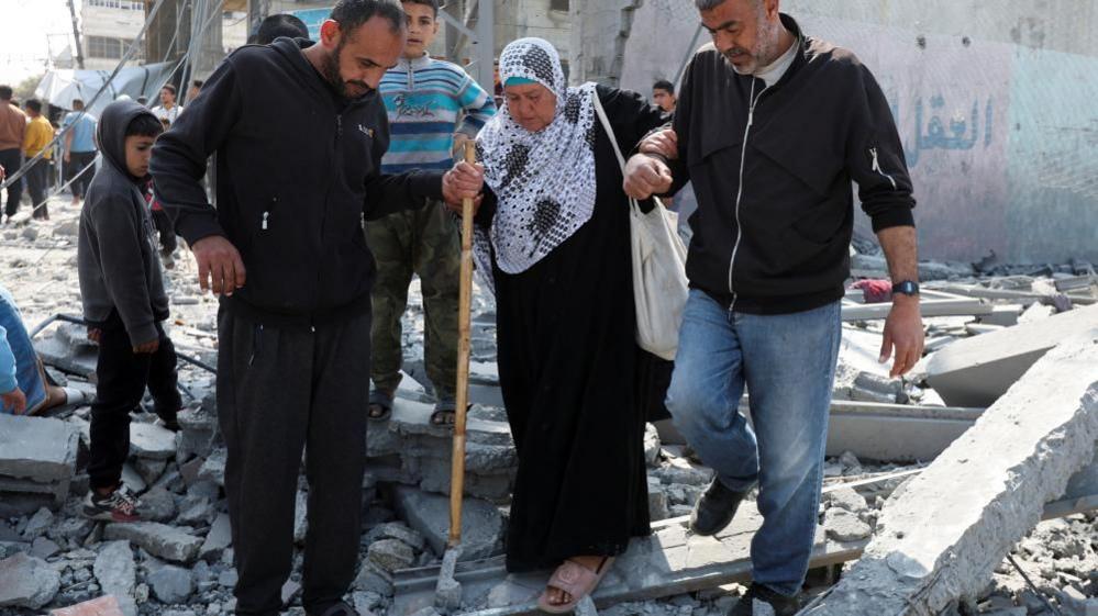 Palestinians walk at the site of an Israeli strike on a residential building, in Deir Al-Balah in the central Gaza Strip (18/03/25)
