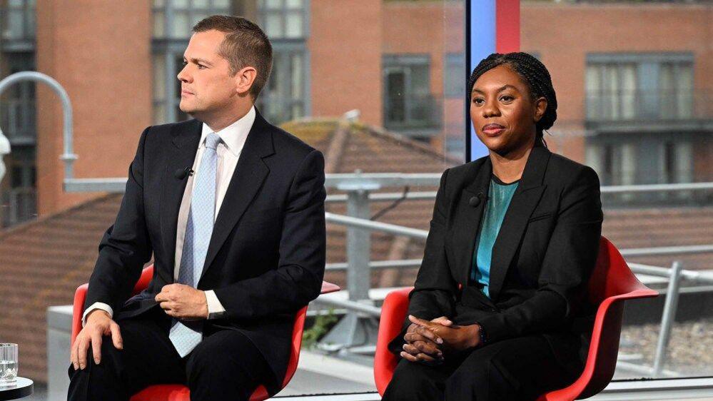 Robert Jenrick MP and Kemi Badenoch MP on the BBC current affairs programme, Sunday with Laura Kuenssberg. They both wear black suits and sit on red chairs in front of a large window.