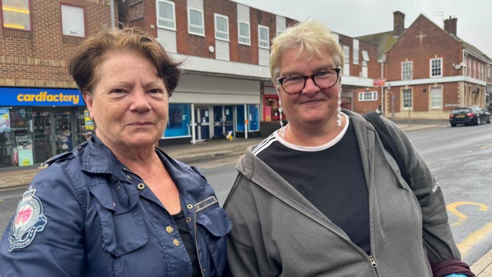 Cynthia has light brunette hair and is wearing a blue jacket with a badge on the shoulder, and pockets and fasteners on the front. Michelle is on the right, and is wearing a black T-shirt with white trim, and a grey zipped hoodie. She is wearing glasses and long white earrings, and has blonde hair. They are standing on Gorleston High Street, with buildings behind them.
