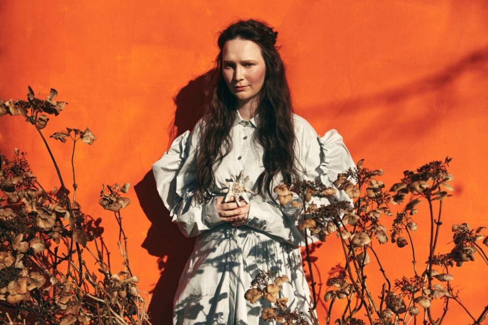 Swiss Eurovision entry Zoë Më stands in a posed mage in a white dress with an orange background, surrounded by flowers.
