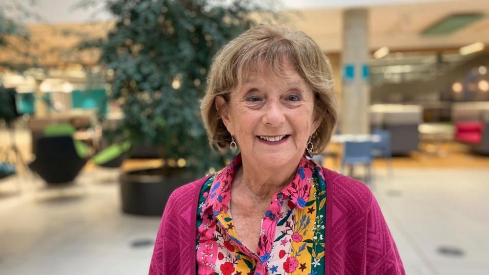 Fiona Brown with medium-length fair hair, smiling and wearing a patterned scarf over a pink sweater. She is in a large building with tables and chairs in the background.