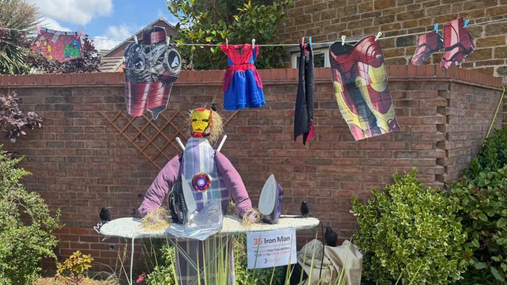 An scarecrow decorated as Iron Man - with a logo on his chest - is leaning on an ironing board with his armour hanging on a washing line. A large brick wall is behind the scarecrow.