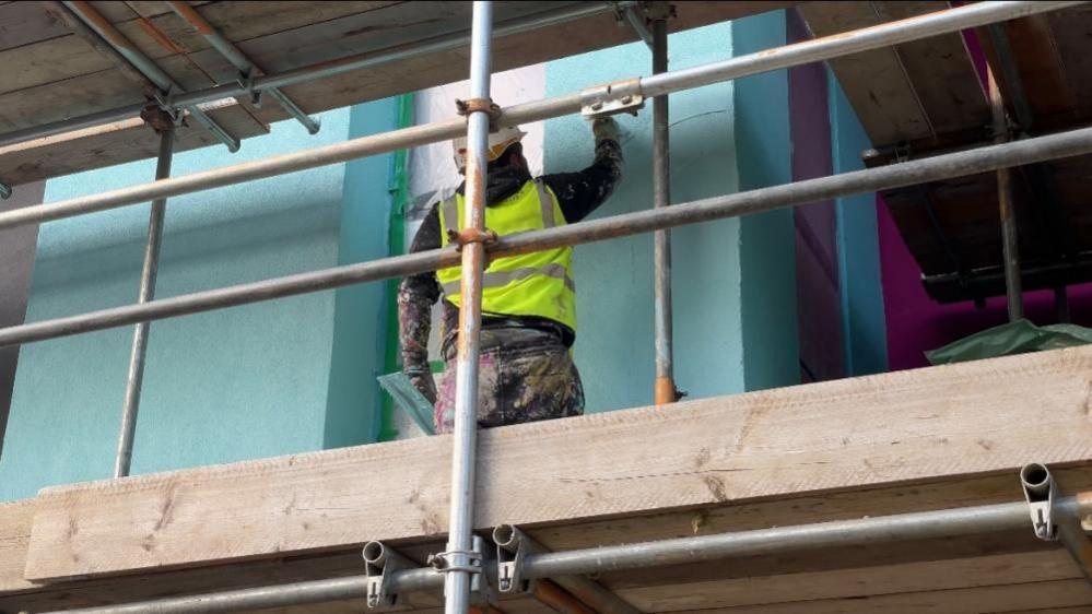 A close up shot of a man wearing a high vis jacket and hard hat, painting the exterior of the centre. The paint is blue. He is standing on scaffolding. 

