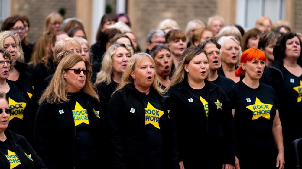 A group of women wearing black T-shirts with yellow Rock Choir T-shirts. They are singing outside a stone building.  One is wearing sunglasses while another has orange hair.