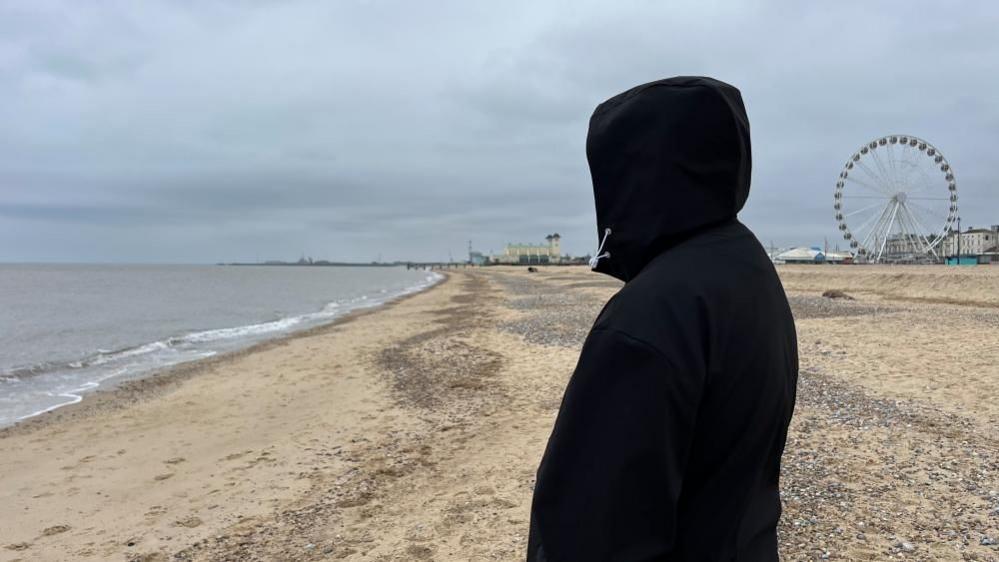 A picture of Leon in a black hoodie, with the hood covering his face to protect his identity. He is pictured on Great Yarmouth's central beach, with the Wellington Pier and big wheel visible in the distance.