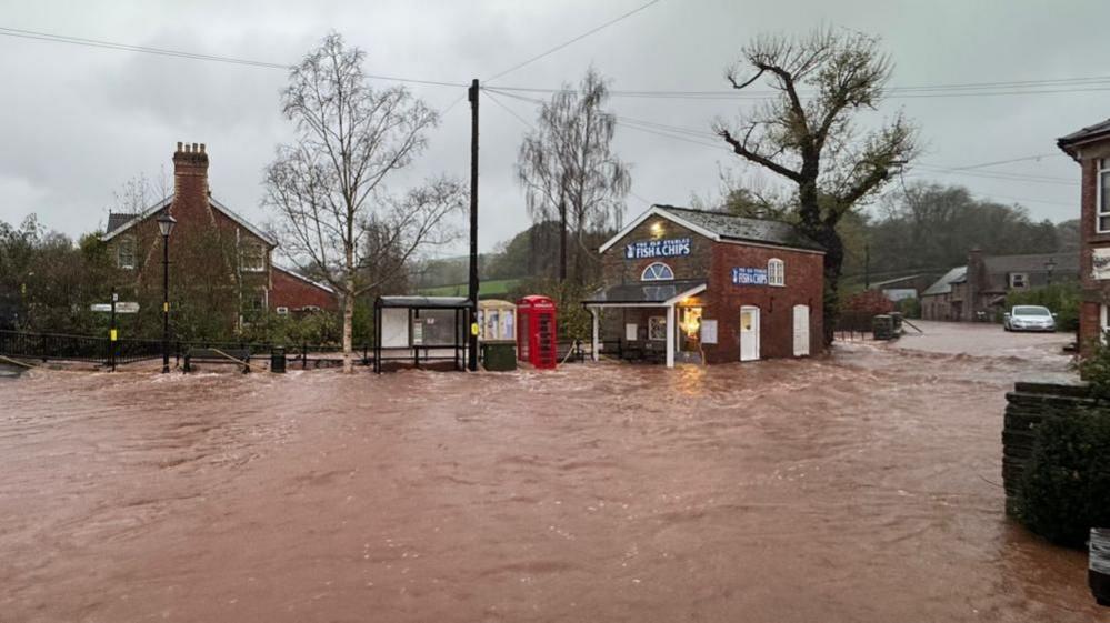 A village that is flooded. Dark orange water covers the road and has submerged parts of a bus stop and telephone box. The water is covering the entrance to a fish and chip shop.
