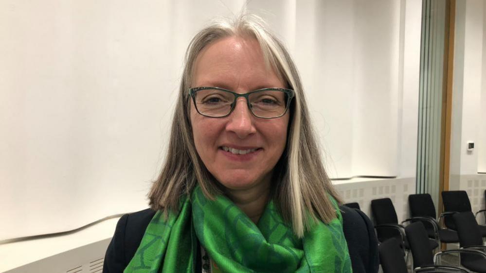 Caroline Topping is facing the camera and smiling she is standing in a council chamber in Suffolk. She is wearing a green scarf. 