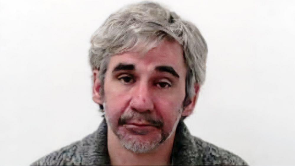 A mugshot of a man with long grey hair and dark eyebrows. He is wearing a grey knitted top.