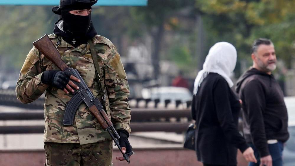 man in camouflage with a gun stands in Damascus as a couple walk past in the background