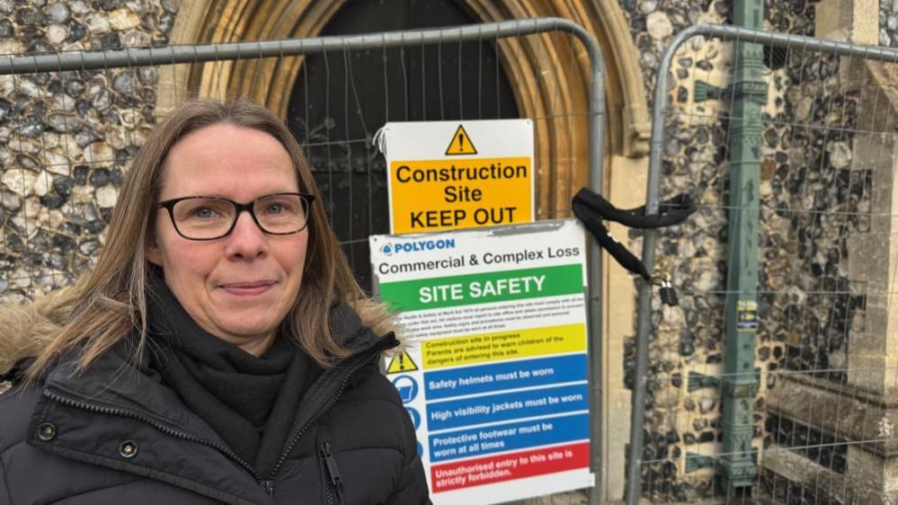 Helen Orford stands outside the entrance of St John's Church, where signage warns of the dangers inside. There is temporary fencing around the building. Ms Orford is wearing a black scarf and a black coat, with a faux fur hood lining. She is wearing black rimmed glasses and has long blonde hair.