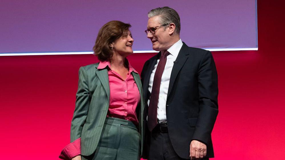 Eluned Morgan wearing a grey suit and pink shirt has her arm around Keir Starmer who is wearing a dark suit, white shirt and a dark red tie. They are smiling at one another