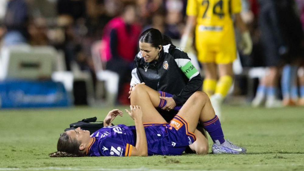 Megan Wynne is lying on the ground with her leg held up by a physiotherapist. She is wearing purple football kit with orange stripes on her sleeve. The physio kneels next to her and is a black and white coat. Blurred behind is a player in a yellow kit.