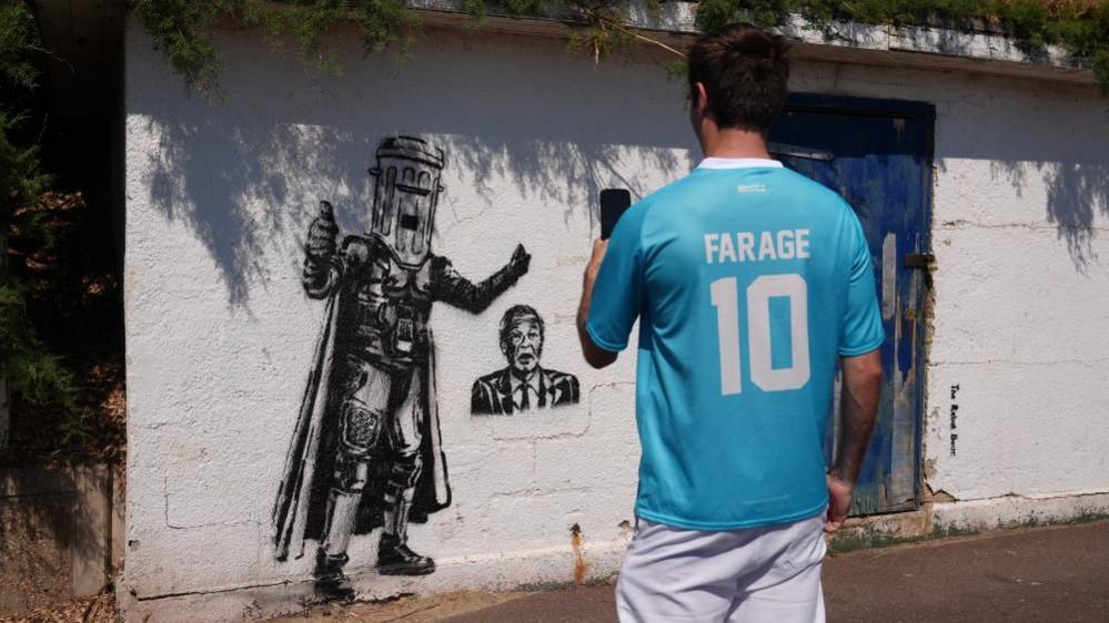 A man wearing a turquoise-coloured shirt with "Farage 10" written in white text on the back is taking a photo on his phone of a mural. The mural is on a white wall and includes two images in black paint. One is of Count Binface, a character wearing a cape and a bin on his head, and the other shows the head and shoulders of Nigel Farage.