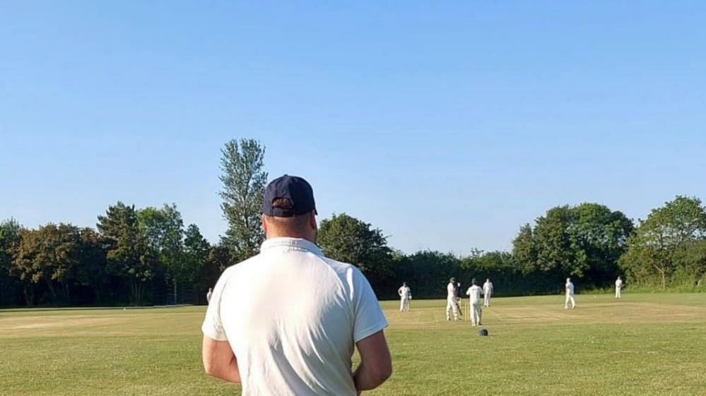 A rear view of a cricketer watching a cricket game being played. A batsman wearing a blue helmet is preparing to face a delivery.