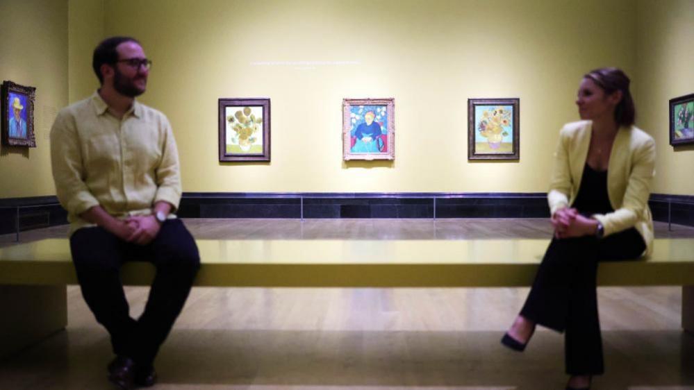 A man and a woman sit on a bench at the National Gallery's Van Gogh exhibition with his paintings in the background