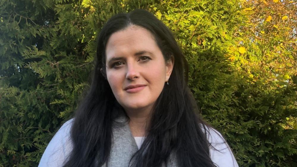 A headshot of Claire Sugden. She has long black hair and is wearing a white t-shirt and a grey scarf. 