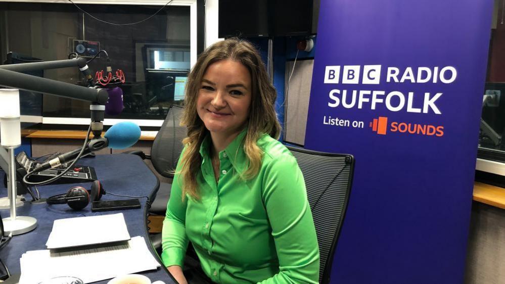 A woman is sitting in the radio studio at BBC Radio Suffolk. She has long hair and is wearing a bright green shirt. 