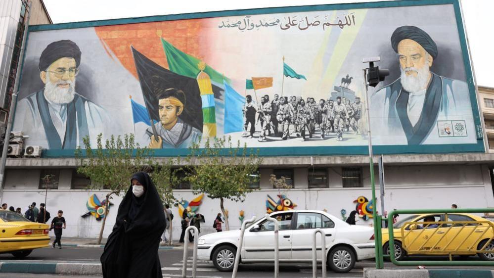 An Iranian woman walks past a wall painting of Iranian Supreme Leader Ayatollah Ali Khamenei and former Iranian Supreme Leader Ayatollah Ruhollah Khomeini in Tehran (21 October 2024)