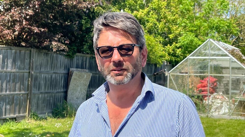 Chris Duncan with dark hair and beard wearing a blue stripped shirt and standing in a garden