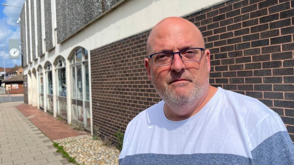 Ian Measures is wearing a blue top, with lighter and darker shades in horizontal bands. He has a shaved head, a close cropped beard and moustache, and is wearing glasses. He is standing by Gorleston Library. The building is a 1970s design, featuring brown bricks, concrete pillars and arches, several infilled with aluminium-framed windows, and flint-faced concrete panels on the upper storey.