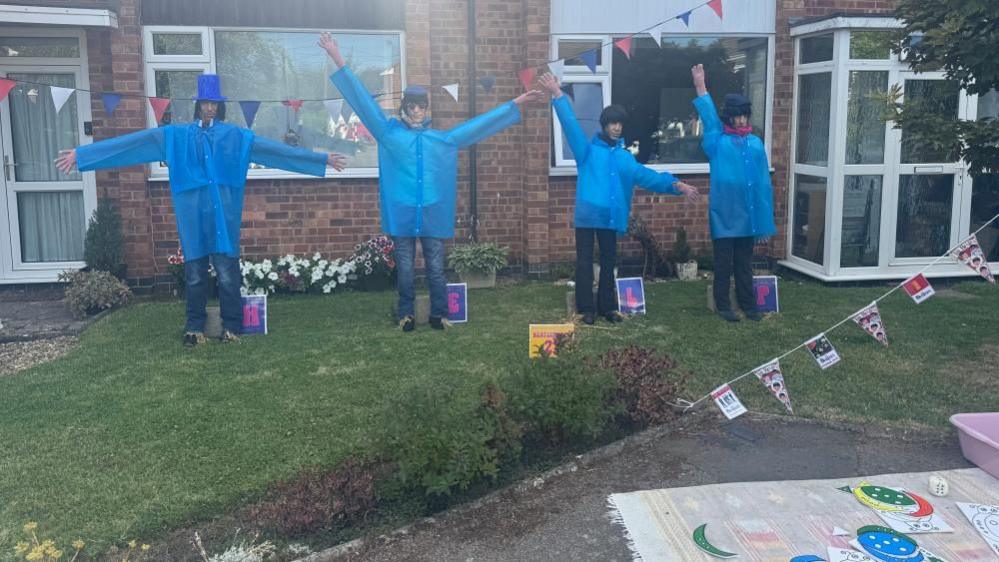 Four scarecrows dressed in bright blue rain coats and jeans on display in a garden. Bunting is hanging to the right. The fronts of two brick houses are behind the scarecrows.