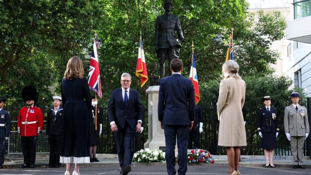 Victoria Starmer, Emmanuel and Brigitte Macron stand with their backs facing the camera, looking at the statue of de Gaulle, which has two wreaths at its base. Keir Starmer walks back towards them