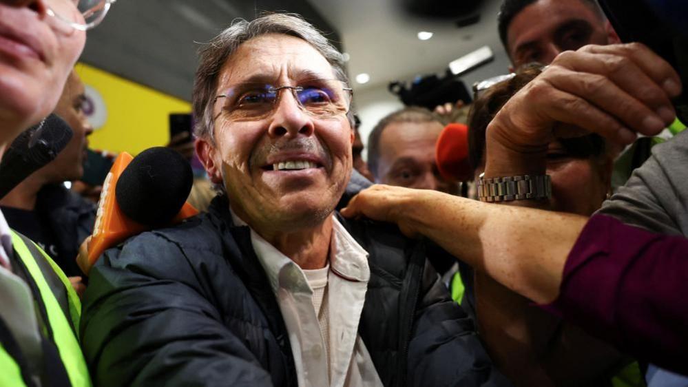 Fabio Ochoa in blue jacket and open-necked white shirt, flanked by a crowd of reporters, smiles as someone out of frame holds his shoulder