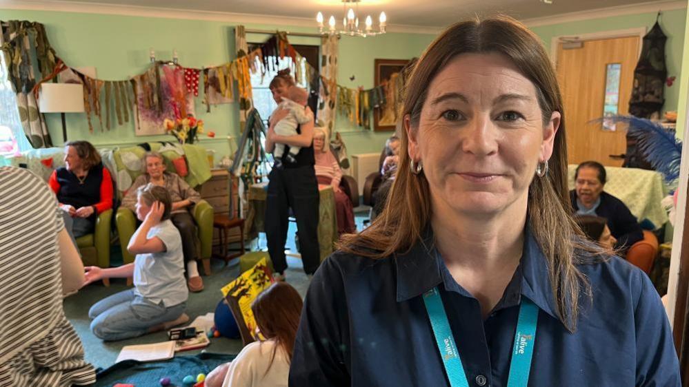 Isobel Jones of the Alive Appeal, who is wearing a blue t-shirt and a light blue lanyard. She is standing in front of a room full of mothers, care home residents, and babies