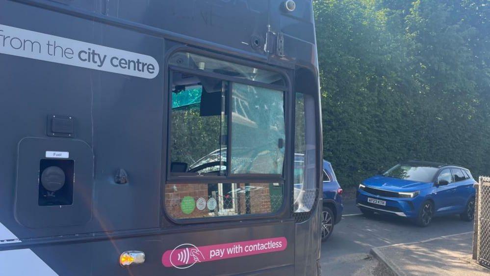 A double decker bus in a dark grey colour, with a pink banner stating: "Pay with contactless" and another banner stating: "from the city centre". The image shows the shattered sliding window of the drivers cab, and other vehicles on the road.