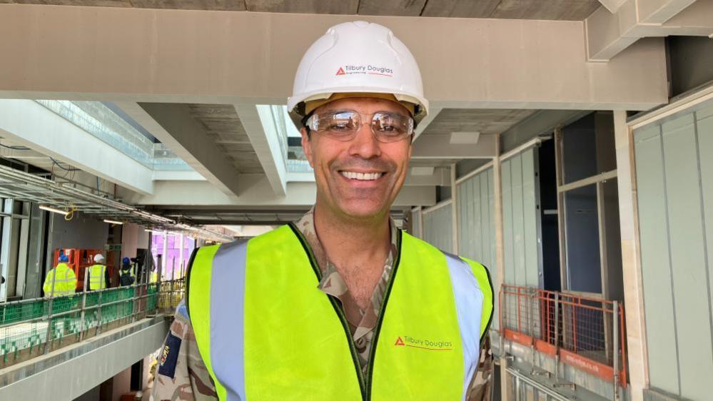 A man wearing military uniform, a bright green safety vest, clear safety goggles and a white hard hat stands in front a construction site.