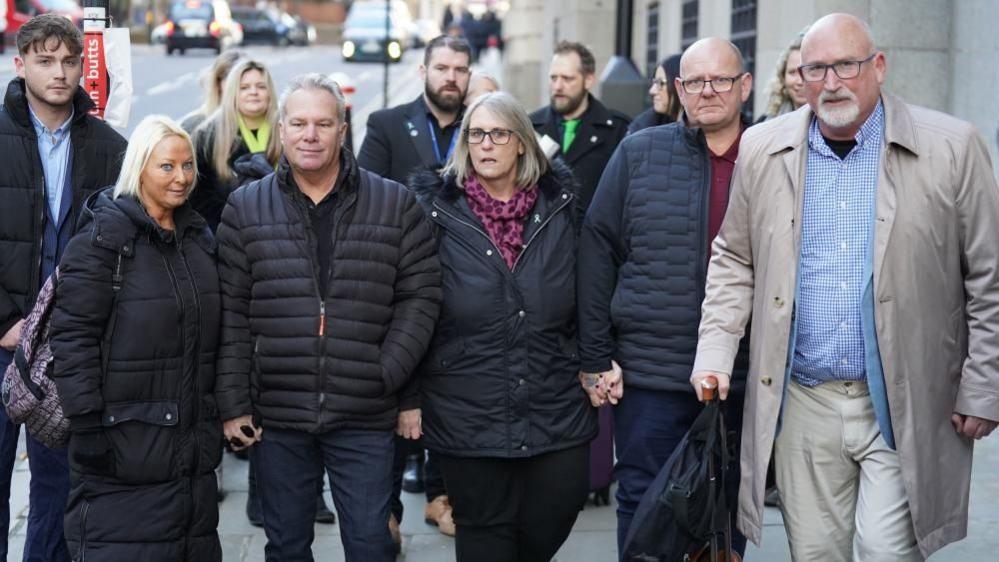 Radd Seiger, with short white hair and beard wearing glasses and a long beige raincoat over white trousers and a blue shirt, walking with two men and two women who are all wearing black coats, on a pavement in a busy city.