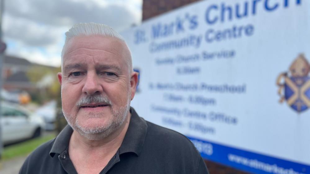 Councillor and vicar Charles Royden, standing outside a sign for St Mark's Church, Bedford. He has grey hair, a bear and is wearing a dark polo style T-shirt. The sign is blurred behind him. 