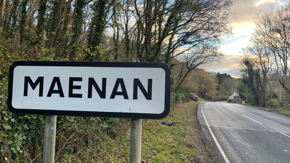 Road sign for the village of Maena, with a country road surrounded by trees